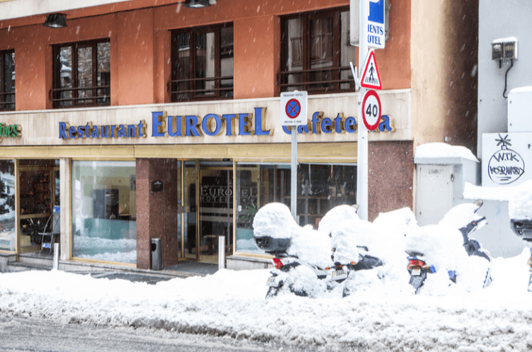 Fachada del Restaurante Cafetería Eurotel durante una intensa nevada. En primer plano, varias motocicletas estacionadas están completamente cubiertas por una gruesa capa de nieve blanca, con señales de tráfico y la entrada acristalada del establecimiento al fondo.
