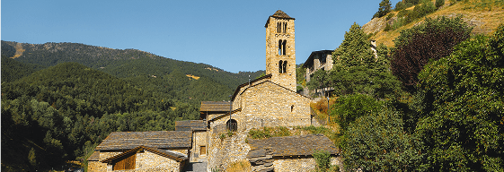 Una església històrica de pedra amb un campanar quadrat alt es troba entre altres edificis de pedra en un paisatge muntanyós i boscós sota un cel blau clar.