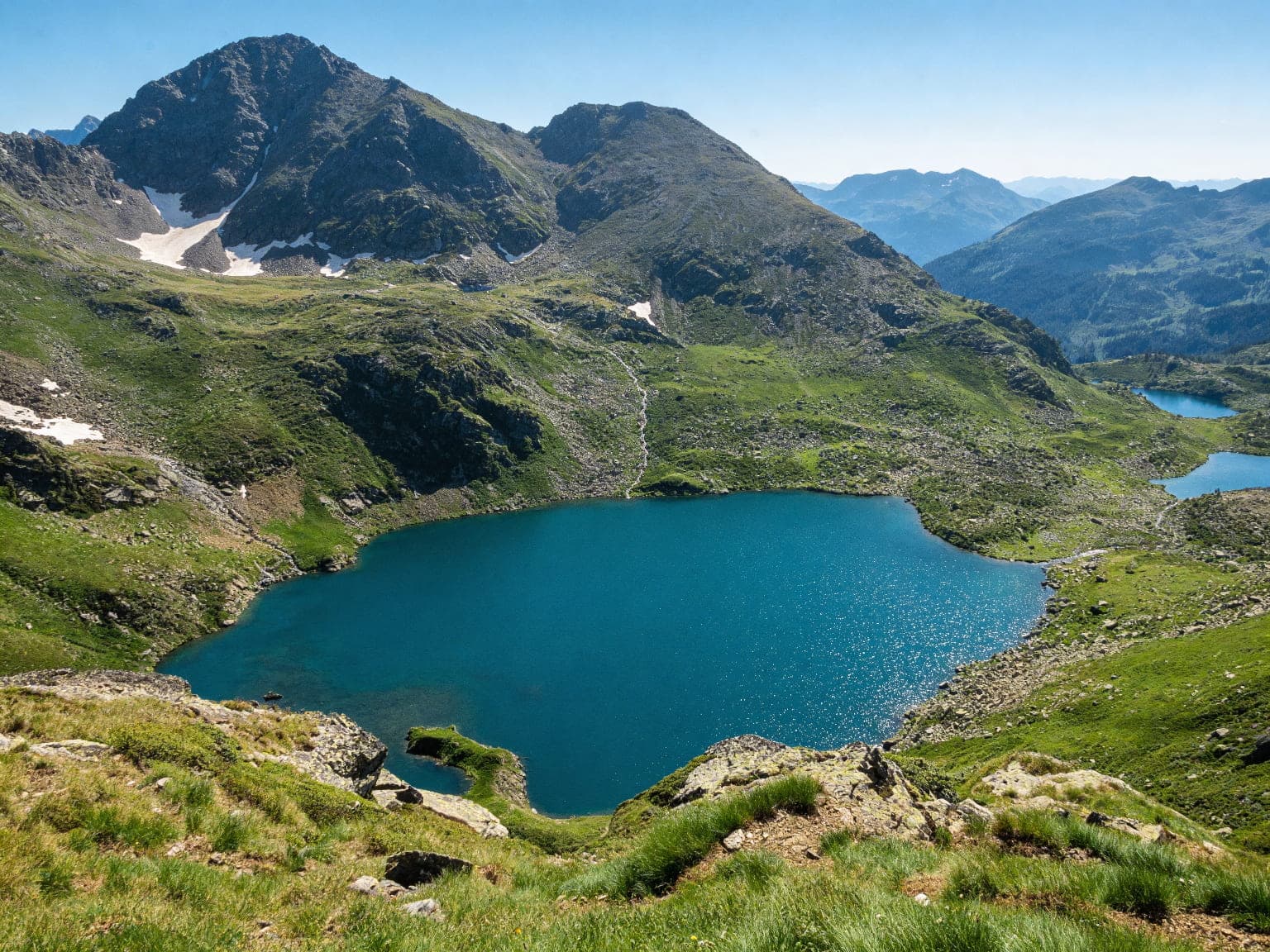 Una vista aèria mostra un llac alpí de color blau vibrant encaixat entre vessants muntanyosos verds i rocosos sota un cel clar. Diversos llacs més petits són visibles a la distància.