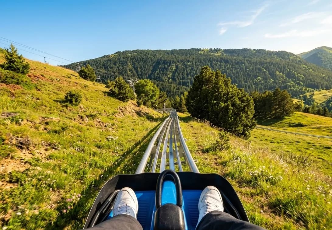 Vista en primera persona des d'un carro de muntanya russa alpina, mostrant les vies que baixen per una muntanya verda i boscosa sota un cel blau clar. Els peus del passatger amb sabatilles blanques són visibles al carro.