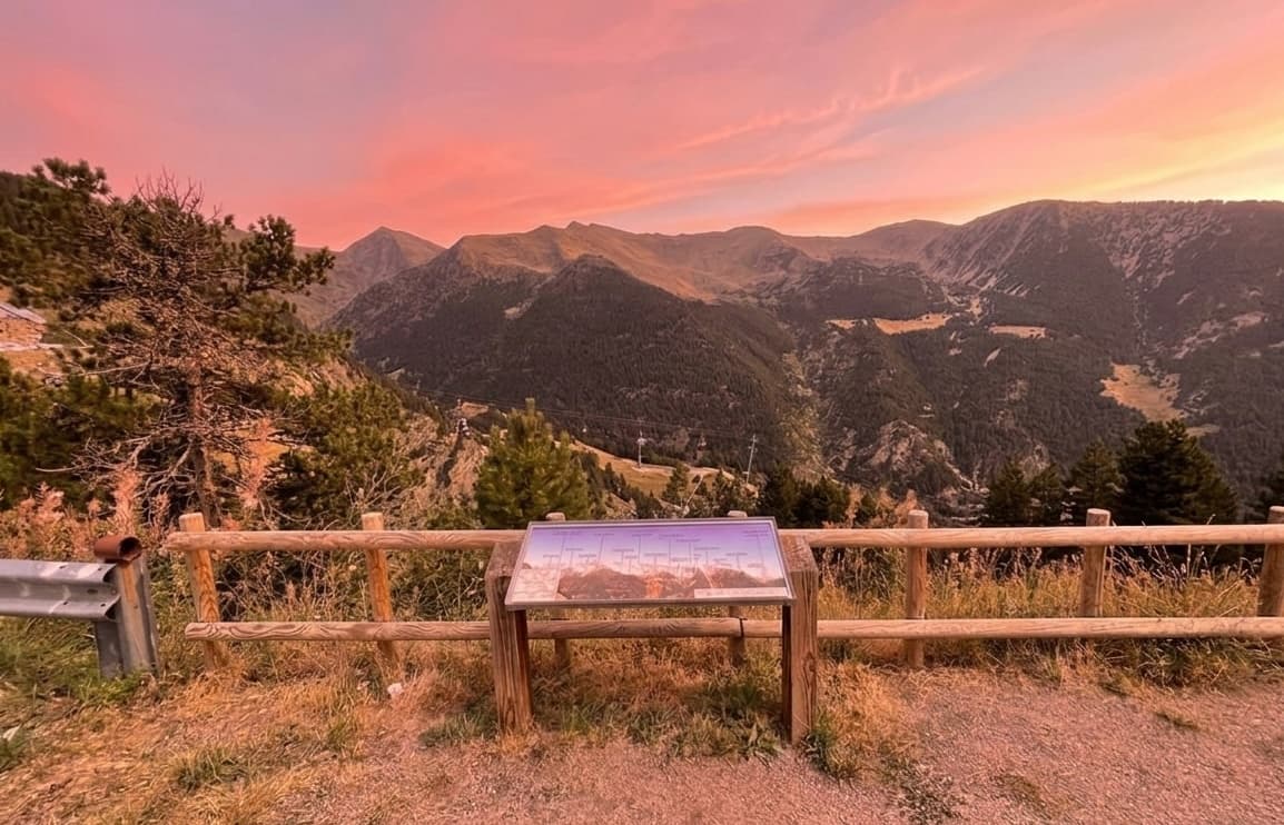 Un panorama muntanyós escènic al capvespre o a la sortida del sol, amb cims escarpats, un bosc dens i un cel vibrant de color rosa i taronja. En primer pla, una tanca de fusta i un cartell informatiu marquen un mirador.