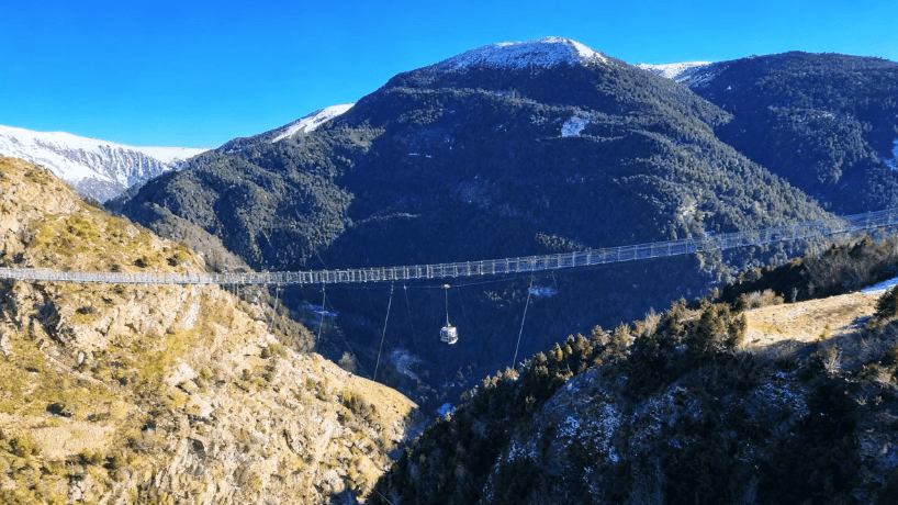 Un largo y moderno puente colgante cruza un profundo valle montañoso cubierto de bosques bajo un cielo azul claro. Una pequeña góndola cerrada cuelga suspendida por cables debajo del puente.