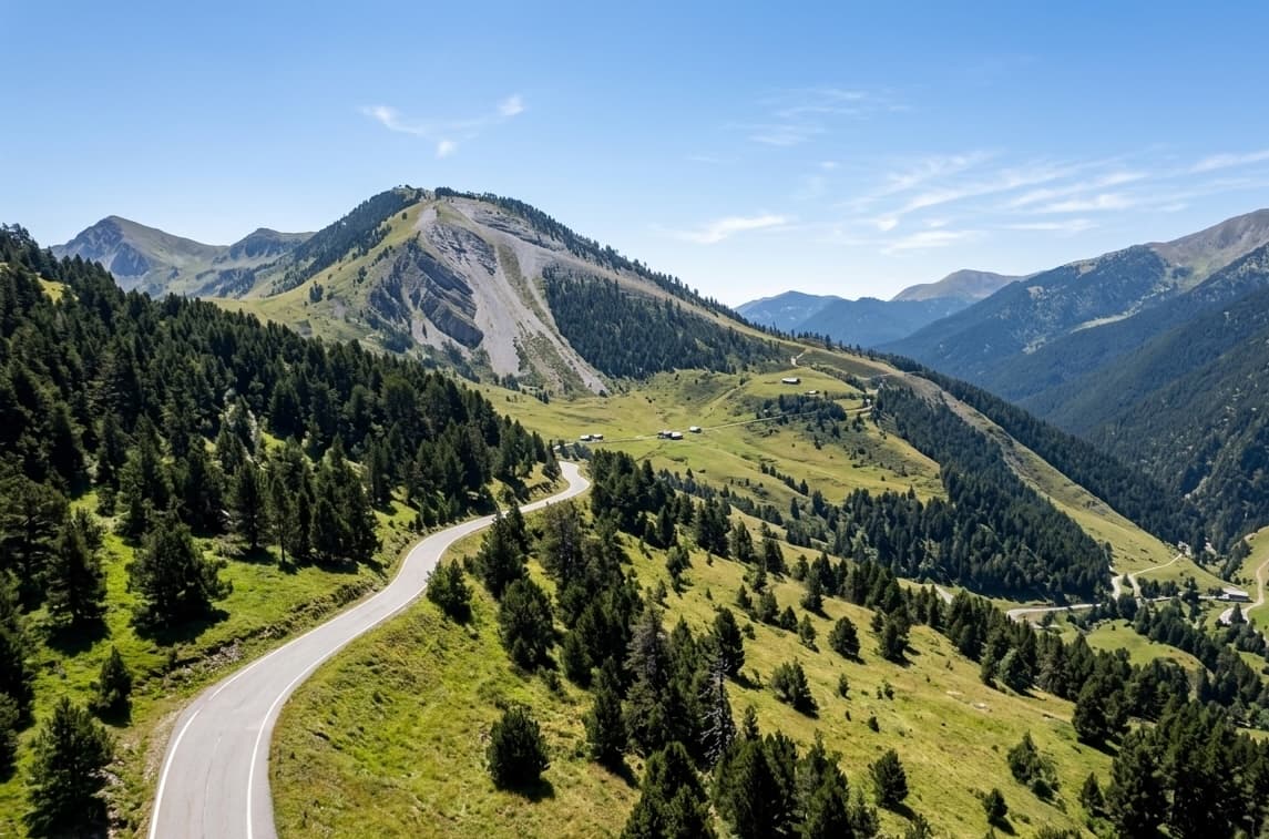 Vista aèria d'una carretera sinuosa que travessa un exuberant paisatge muntanyós verd amb densos boscos de pins sota un cel blau clar. Es veuen cims i valls distants al fons.