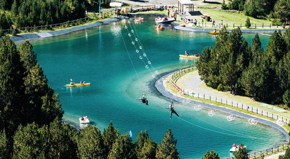 Vista aérea de un parque de aventuras con un lago azul claro. Personas se deslizan en tirolina sobre el agua, mientras otras navegan en kayak y patines de pedales, rodeadas de pinos verdes y un sendero pavimentado.