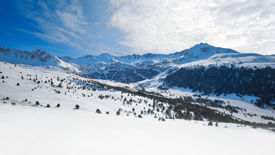 Vista panorámica de una cordillera cubierta de nieve bajo un cielo azul brillante con nubes dispersas. Pinos salpican las laderas nevadas y se ven pistas de esquí a lo lejos.