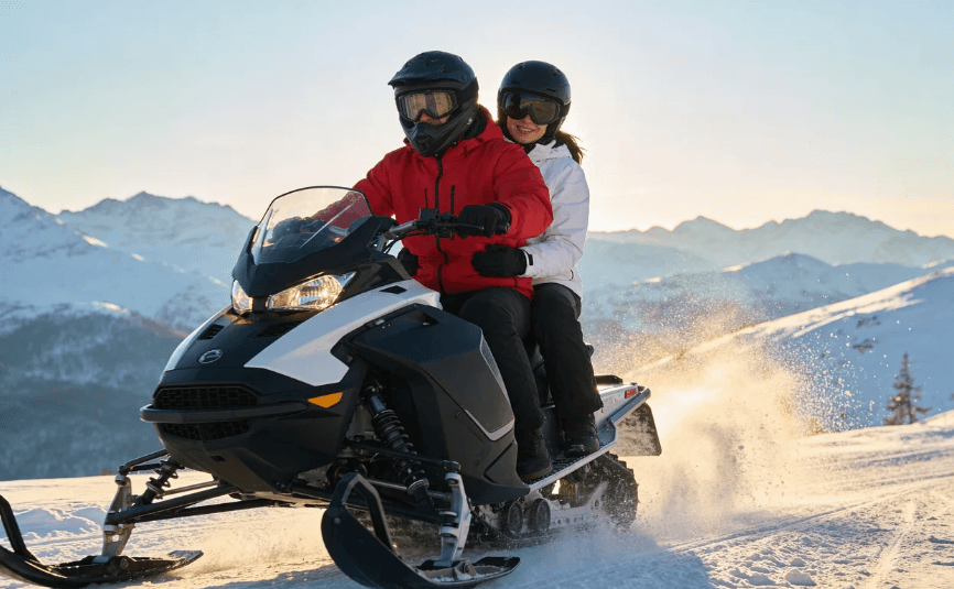 Pareja con cascos protectores conduciendo una moto de nieve plateada por un camino nevado y sinuoso, rodeados de pinos y un paisaje invernal bajo un cielo nublado.