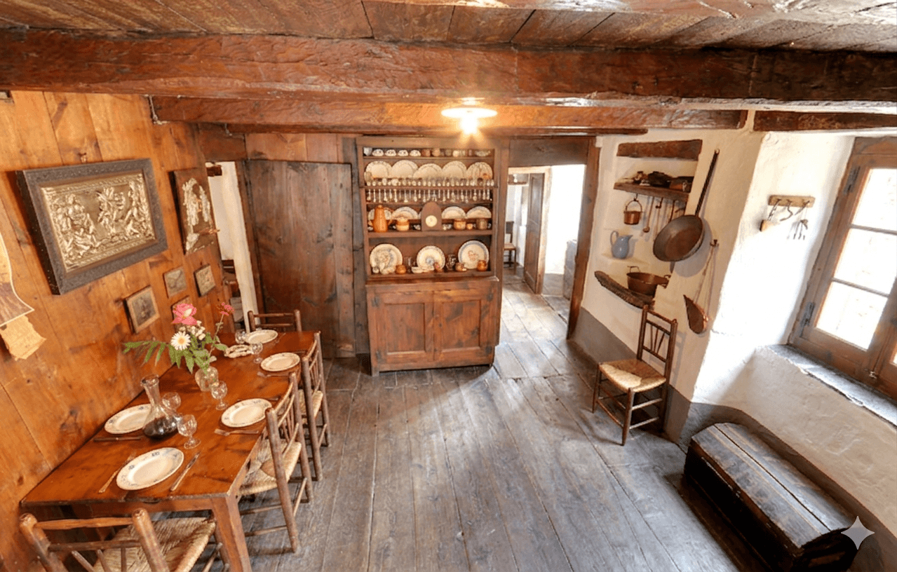 Una habitación rústica y tradicional presenta una larga mesa de comedor de madera puesta con platos y vasos, junto a un aparador que exhibe varias vajillas. Vigas y paneles de madera expuestos crean una atmósfera cálida e histórica, con utensilios de cocina colgando en una pared blanca cercana.