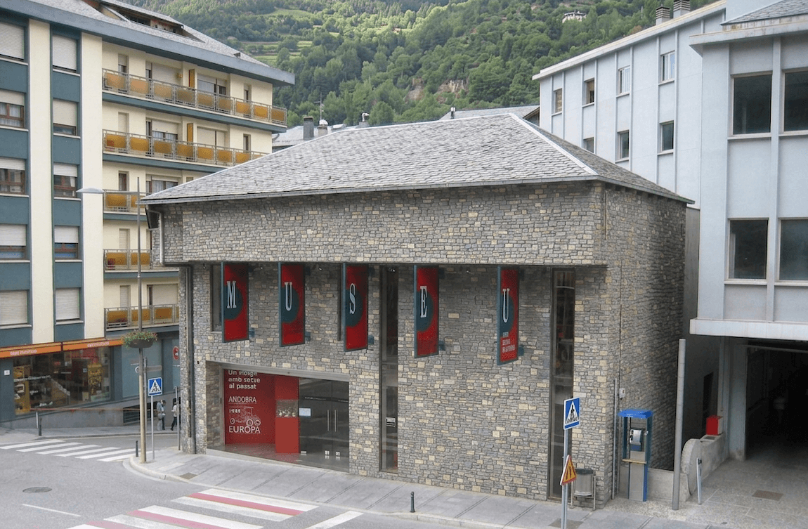 Un edifici amb façana de pedra i teulada de teules grises, amb pancartes vermelles que formen 'MUSEU' a la seva façana, situat en una cantonada amb un pas de vianants. Muntanyes verdes s'eleven al fons darrere d'edificis moderns circumdants.