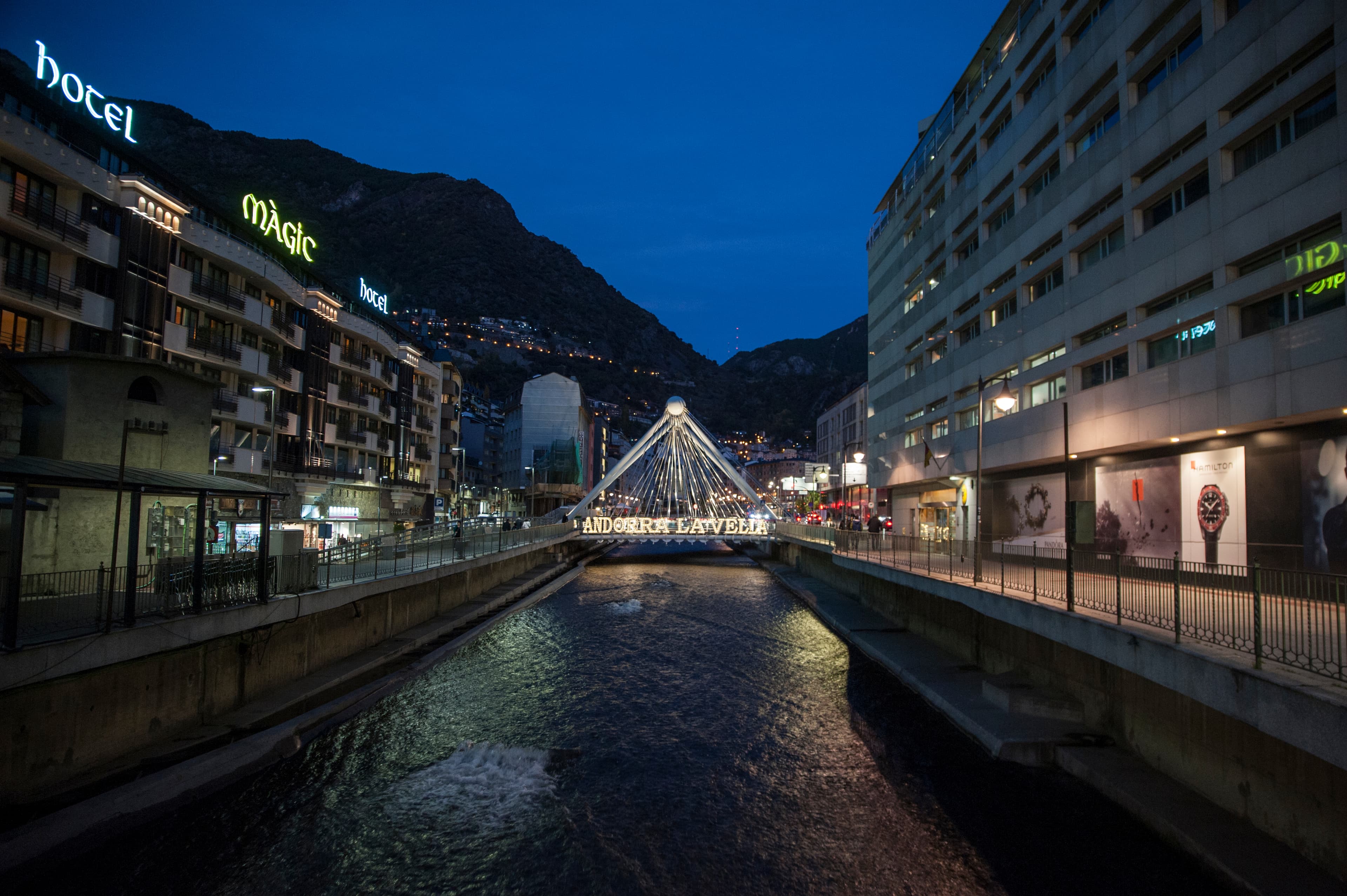 Una vista nocturna d'Andorra la Vella, mostrant el Pont de París il·luminat sobre un riu, flanquejat per edificis amb rètols lluminosos i muntanyes al fons.