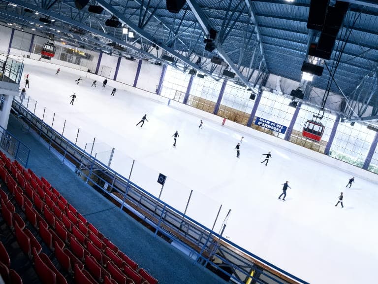 Una pista de hielo cubierta con varias personas patinando, con asientos rojos para espectadores y una cabina de telecabina roja suspendida del techo.