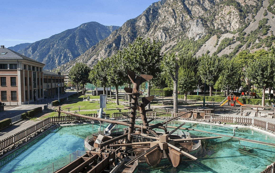 Un parque público con una gran escultura de agua cinética oxidada en una piscina turquesa, rodeado de árboles verdes, un parque infantil y edificios, con majestuosas montañas bajo un cielo azul claro al fondo.