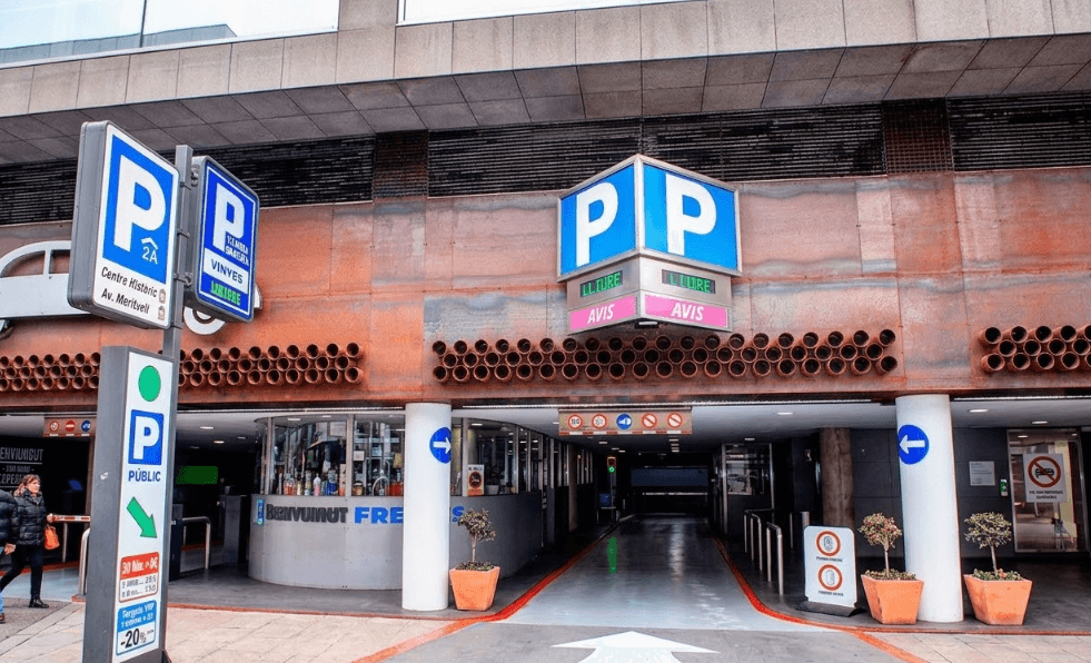 Se muestra la entrada a un aparcamiento subterráneo, con múltiples señales azules de "P". Un gran cartel cúbico sobre la entrada indica "LLTURE" (libre) y "AVIS" (aviso) con luces verdes y rosas, mientras que una señal alta a la izquierda apunta a "Centre Historic" y aparcamiento "Público".