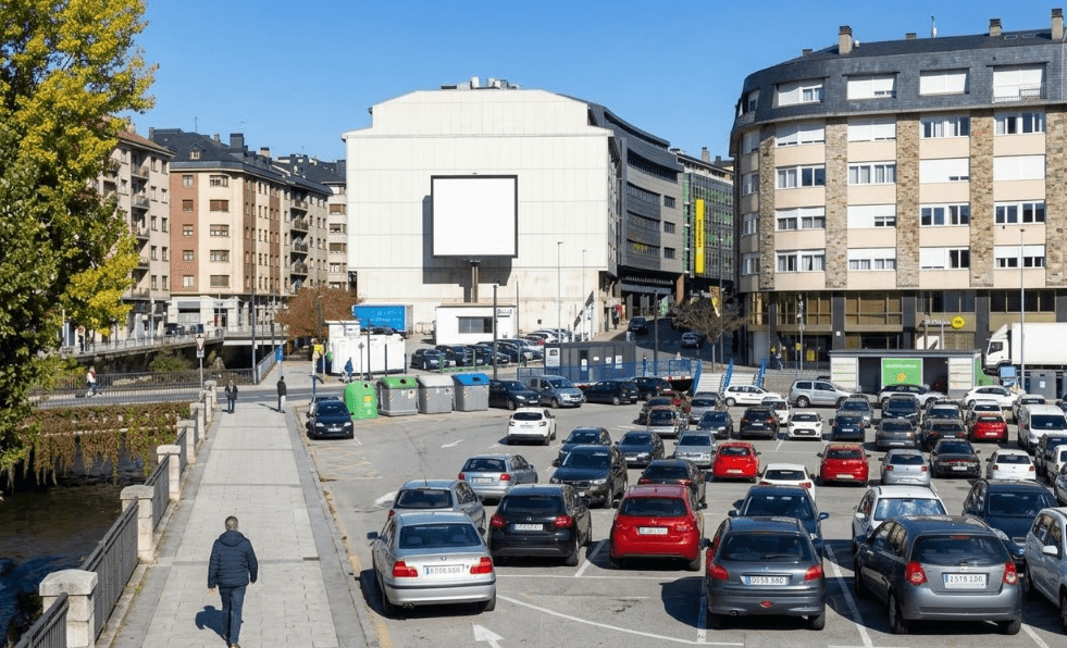 Una escena urbana amb un gran aparcament a l'aire lliure ple de cotxes, vorejat per edificis de diversos pisos i un passeig per a vianants al costat d'un riu. Un edifici prominent al fons té una gran pantalla blanca en blanc.