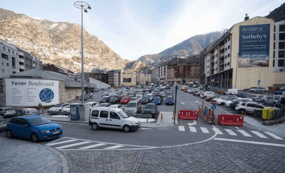 Una vista amplia de un concurrido estacionamiento al aire libre en una ciudad montañosa, con numerosos coches aparcados entre edificios y picos nevados al fondo. Se ven letreros de "Fener Boulevard" y "Andorra Sotheby's International Realty" en edificios adyacentes.