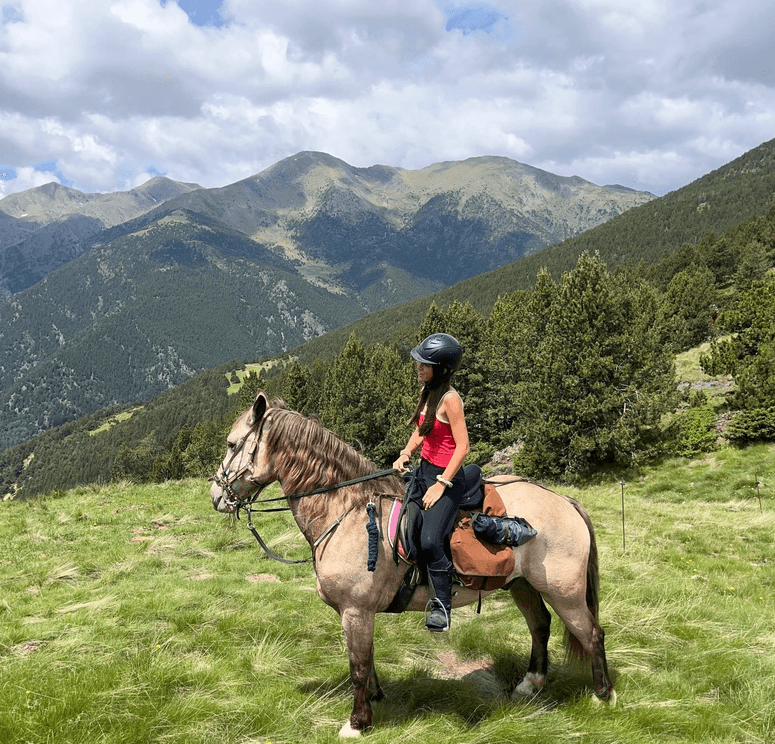 Una persona amb samarreta vermella i casc munta un cavall marró clar en un vessant de muntanya herbós, amb boscos de pins i cims distants sota un cel ennuvolat.