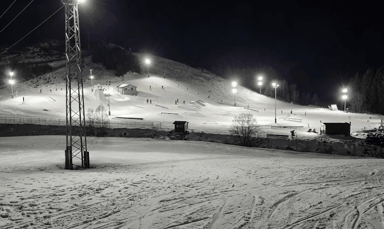 Fotografia nocturna des d'una perspectiva a nivell del sòl a la base d'un snowpark il·luminat. En primer pla s'aprecia la textura de la neu amb petjades d'esquís, juntament amb una torre metàl·lica d'il·luminació a l'esquerra. Al fons, el vessant de la muntanya està equipat amb rampes, rails i focus brillants que destaquen sobre la foscor del cel, amb petites cabanes de fusta i una àrea d'aparcament a la dreta.