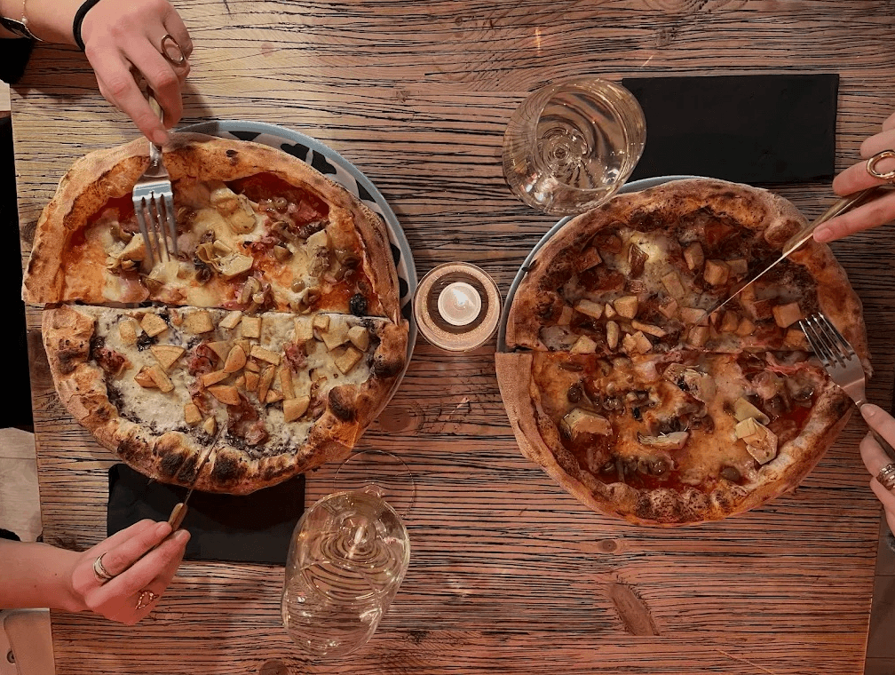 Vista zenital de dues pizzes grans amb diversos ingredients, incloent carxofes i xampinyons, sent tallades i menjades per diverses mans en una taula de fusta rústica, amb begudes i una espelma.
