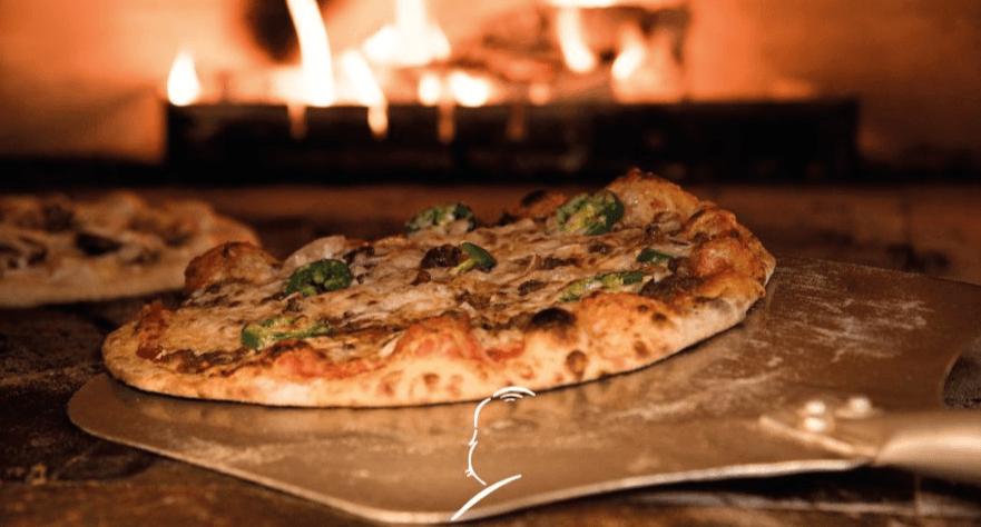 Una pizza acabada de fer amb pebrots verds i formatge reposa sobre una pala de metall, sent introduïda en un forn de llenya incandescent amb flames visibles. Una altra pizza borrosa es veu al fons del forn.