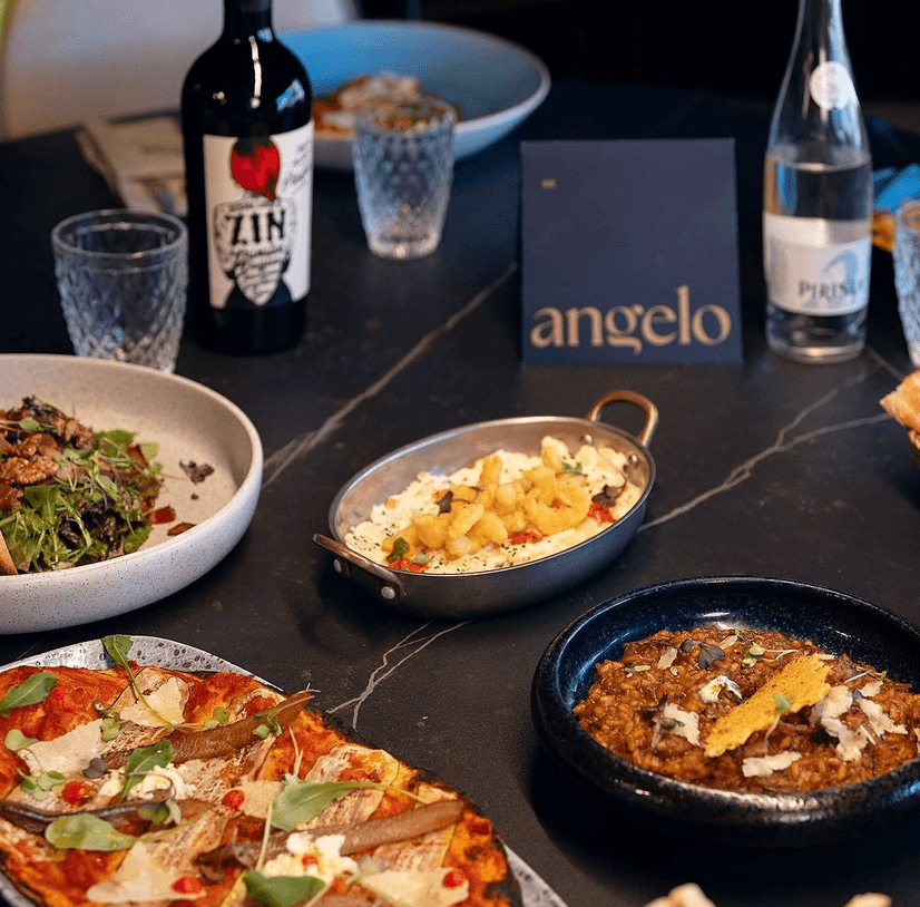 Un primer pla d'una taula de restaurant amb diversos plats, incloent una pizza amb anxoves, una amanida verda, una paella de gambes fregides i un bol de risotto, al costat d'una ampolla de vi negre i una carta de menú.
