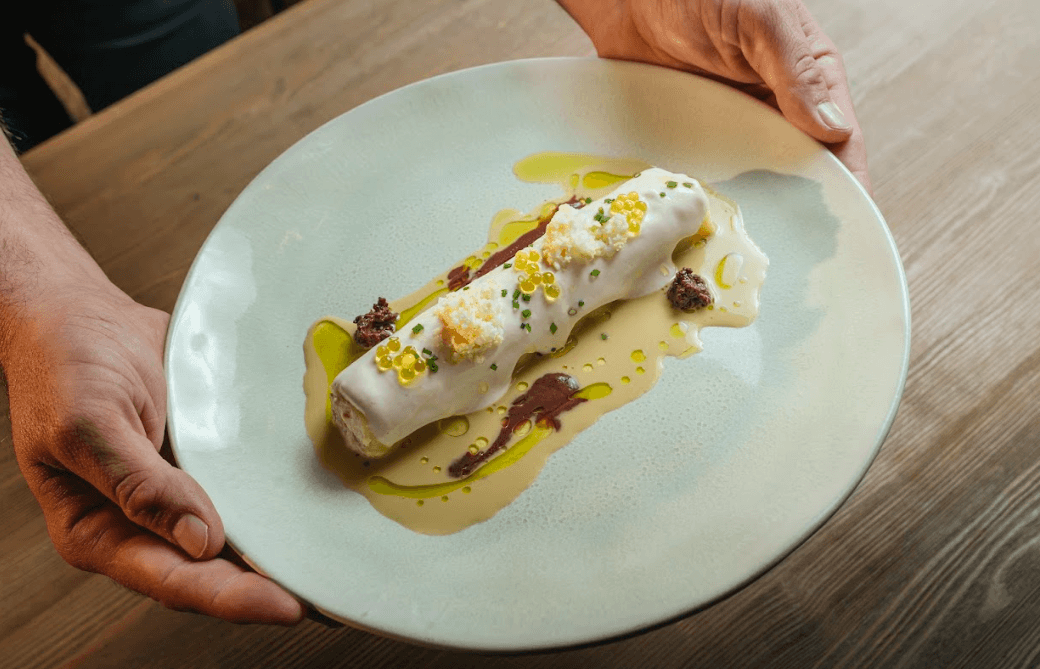 Primer pla de mans sostenint un plat clar amb un plat gurmet. El plat principal és un rotllo tipus caneló cobert amb salsa blanca, guarnit amb esferes grogues, molles blanques, cibulet i tapenade d'olives negres, tot sobre una salsa cremosa amb rajolins d'oli d'oliva.