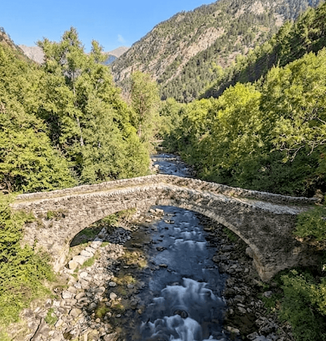 Un antiguo puente de arco de piedra cruza un río rocoso que fluye por un exuberante valle montañoso verde bajo un cielo despejado.