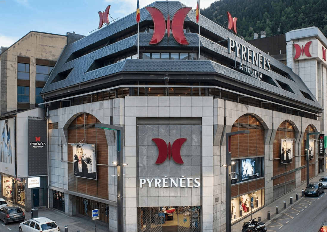 Vista exterior de los grandes almacenes Pyrénées Andorra, un edificio moderno y grande con fachada gris, techo oscuro multifacético y prominentes logotipos rojos de mariposa. La calle de enfrente tiene coches aparcados y se ven montañas al fondo.
