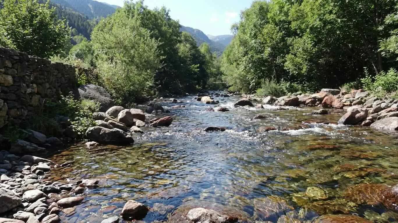 Un riu clar i rocós flueix per una vall verda, flanquejat per arbres densos i un mur de pedra a l'esquerra. Es veuen muntanyes a la distància sota un cel clar.