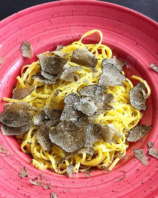 Primer pla de pasta groga, possiblement tagliatelle o linguine, coberta generosament amb fines làmines de tòfona negra, servida en un bol rosa vibrant.