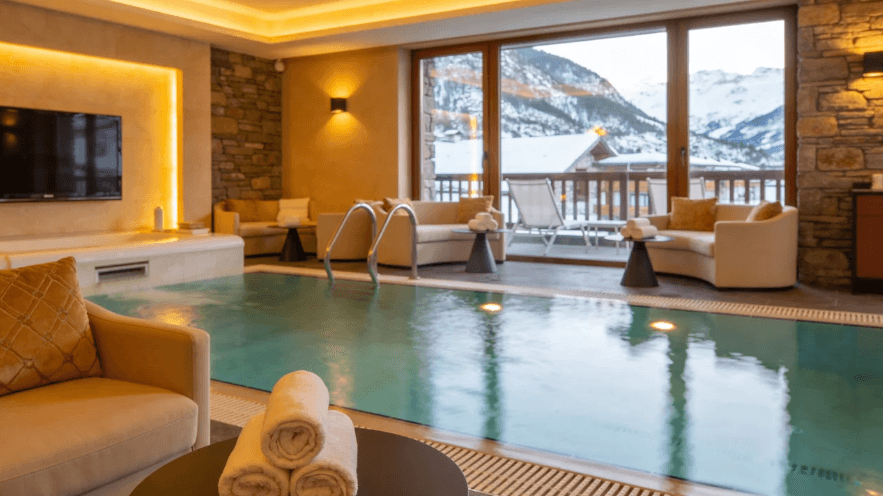 Interior de un lujoso spa con piscina climatizada, sofás de color crema y toallas enrolladas. Al fondo, grandes ventanales ofrecen vistas a un paisaje de montañas nevadas y una terraza exterior.