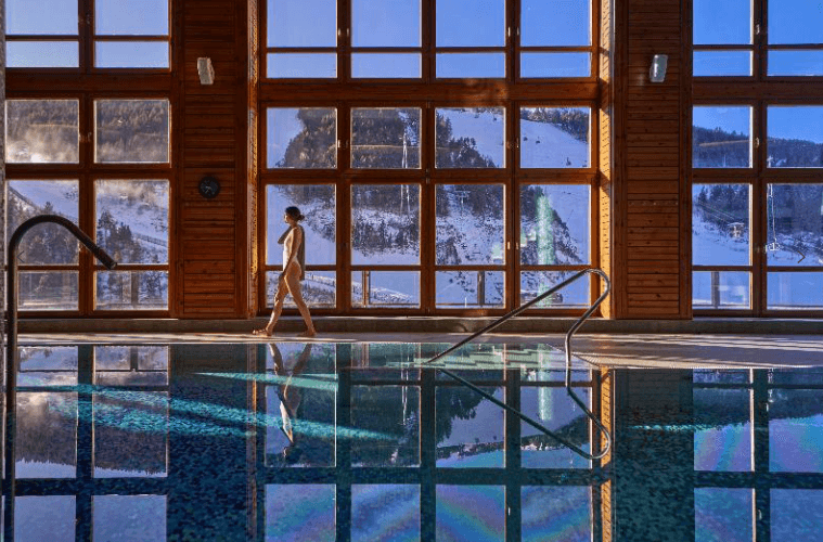 Una dona amb banyador blanc camina per la vora d'una piscina coberta, amb grans finestrals que revelen un paisatge de muntanya nevat a l'exterior.