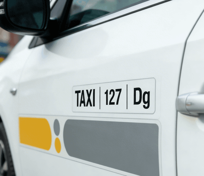 Primer plano del lateral de un taxi blanco con una pegatina que muestra "TAXI | 127 | Dg" y un diseño gráfico amarillo y gris debajo.
