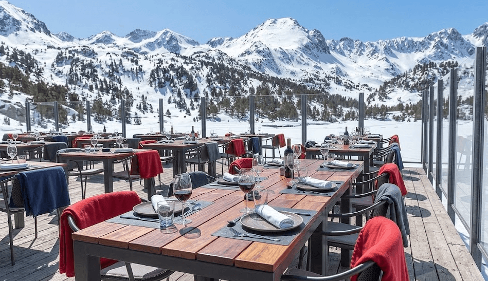 Una terrassa de restaurant amb taules i cadires de fusta, preparades amb parament i copes de vi, amb vistes a una serralada nevada i un llac parcialment glaçat. Mantes vermelles i blaves estan col·locades sobre les cadires.