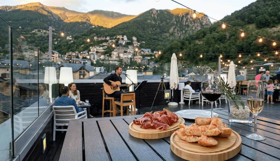 Una animada terrassa al terrat al capvespre, amb un músic tocant la guitarra per als clients que gaudeixen de begudes i aperitius com pernil curat i pa amb tomàquet, amb un pintoresc poble de muntanya al fons.