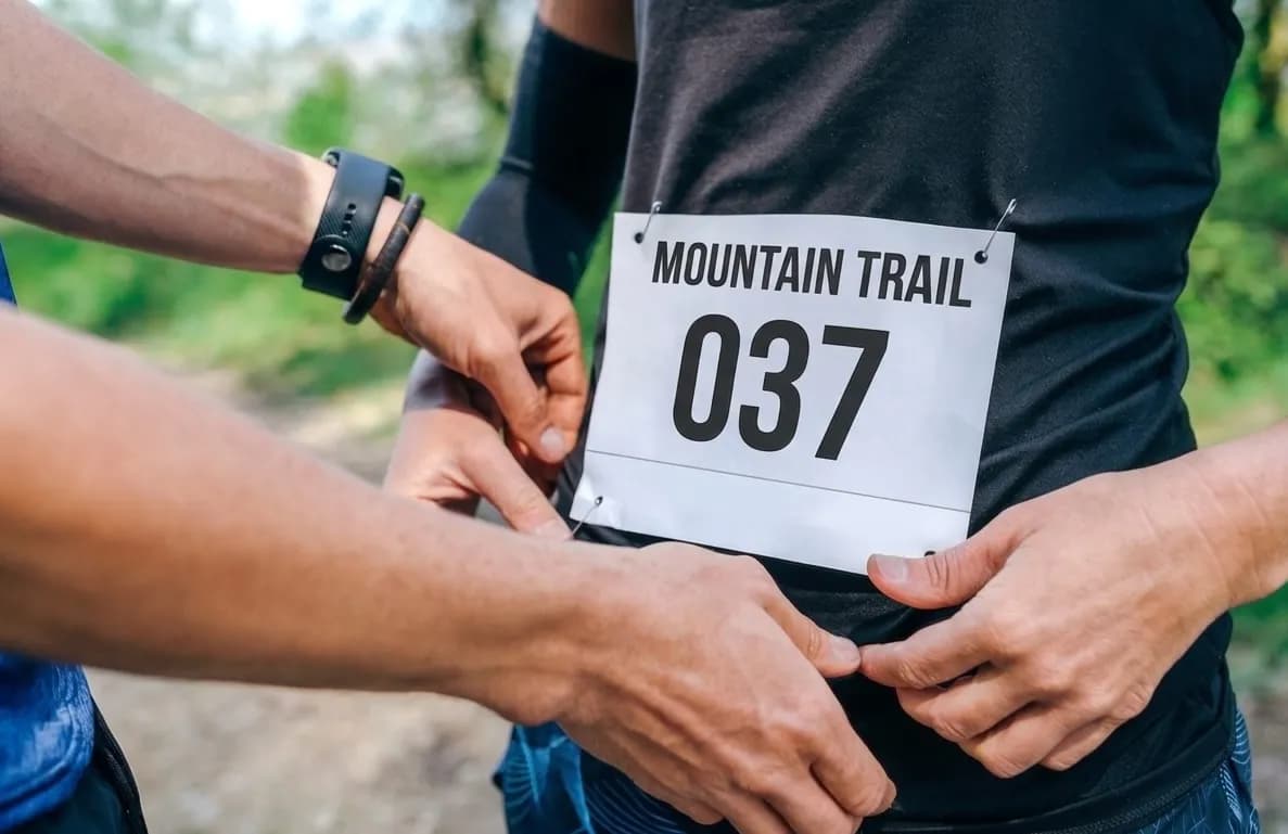 Primer pla de mans subjectant un dorsal blanc amb "MOUNTAIN TRAIL 037" a la samarreta negra d'una persona amb imperdibles. Una persona porta un rellotge intel·ligent.