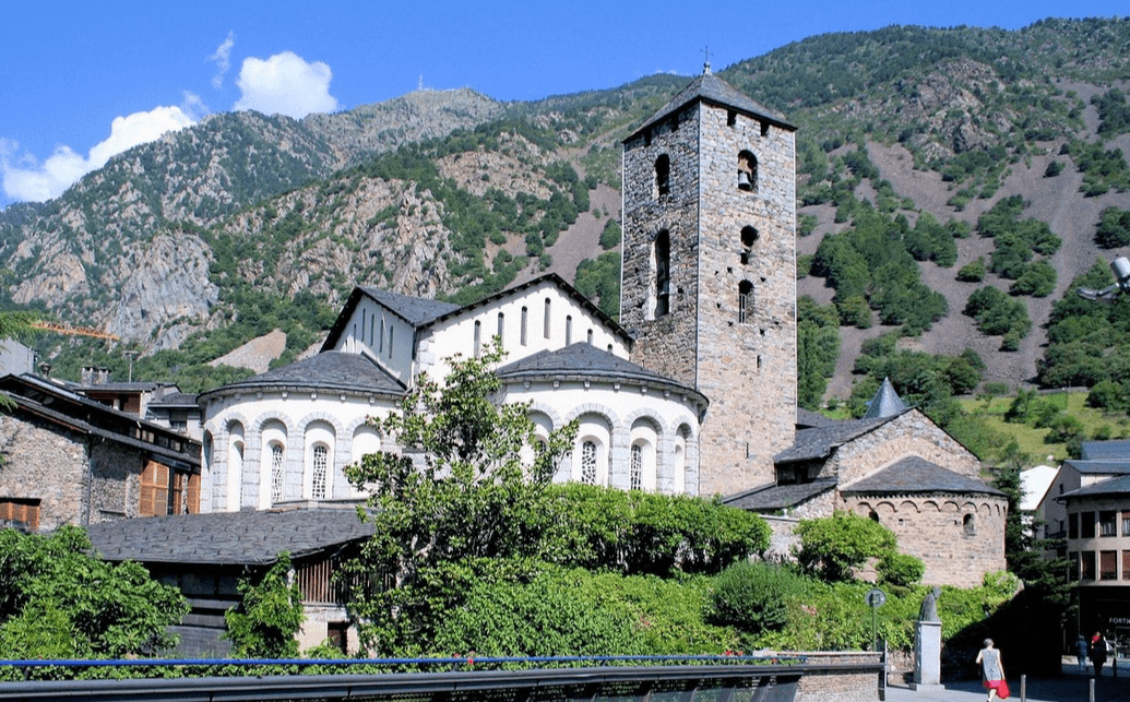Una iglesia histórica de piedra con un alto campanario y ábsides redondeados se encuentra entre árboles verdes y edificios, con un telón de fondo de montañas escarpadas y boscosas bajo un cielo azul.