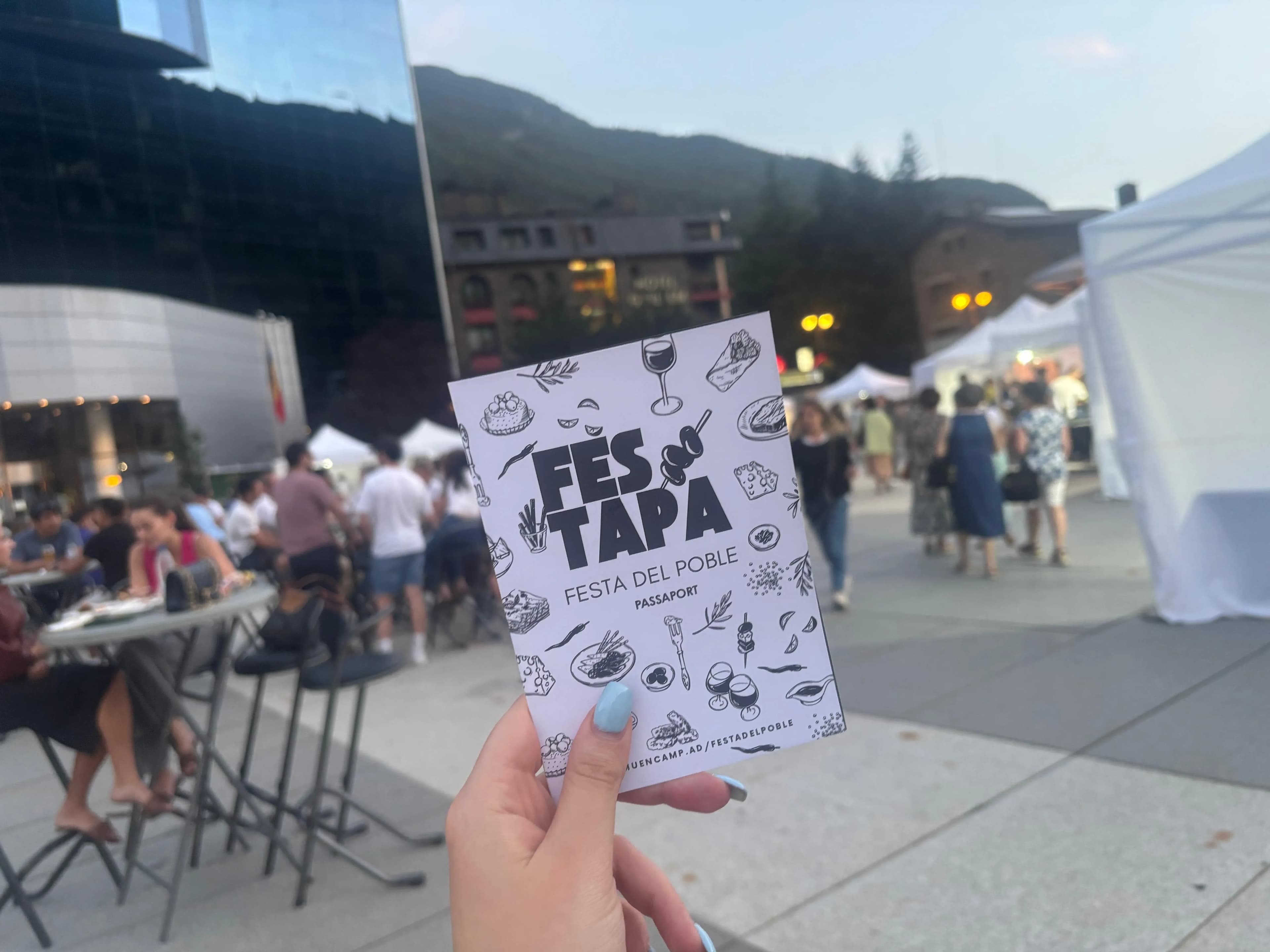 Una mà sosté un fullet blanc de 'FES & TAPA FESTA DEL POBLE PASSAPORT' amb il·lustracions de menjar. Al fons, es veu un festival a l'aire lliure amb gent, taules, carpes blanques, edificis i muntanyes.