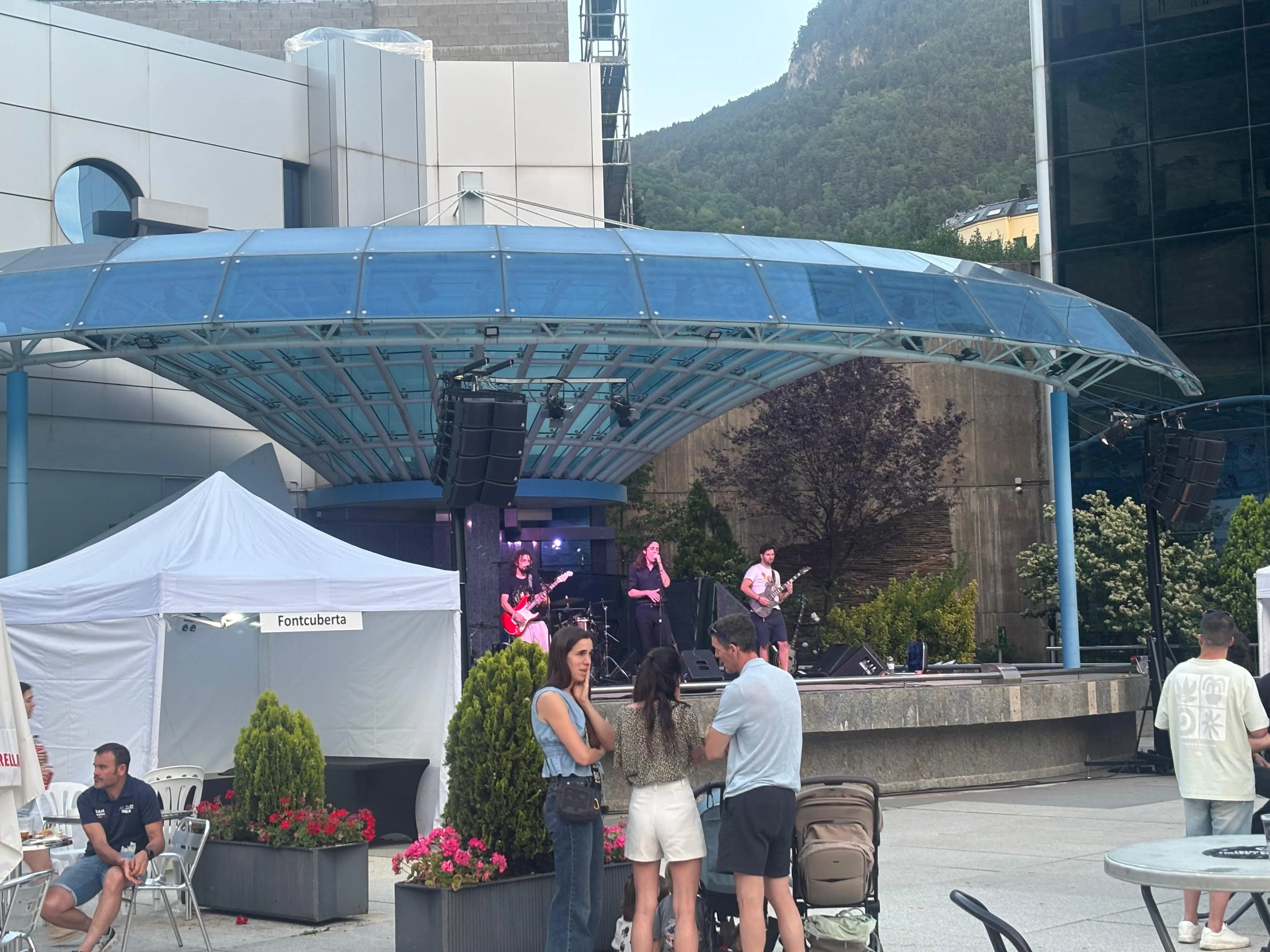 Escena de concert a l'aire lliure amb una banda actuant en un escenari sota un dosser blau. Diverses persones estan reunides en primer pla, amb edificis moderns i una muntanya boscosa al fons.