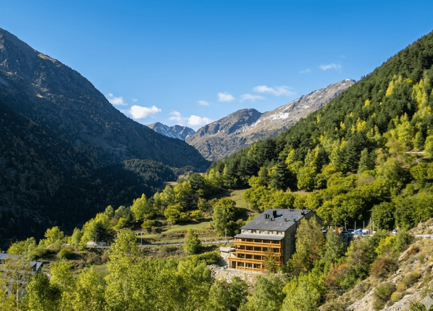 Fotografia en pla general d'un hotel de muntanya amb balcons de fusta, situat al vessant d'una vall frondosa i verda. Al fons, s'observen imponents muntanyes sota un cel blau clar en un dia assolellat.