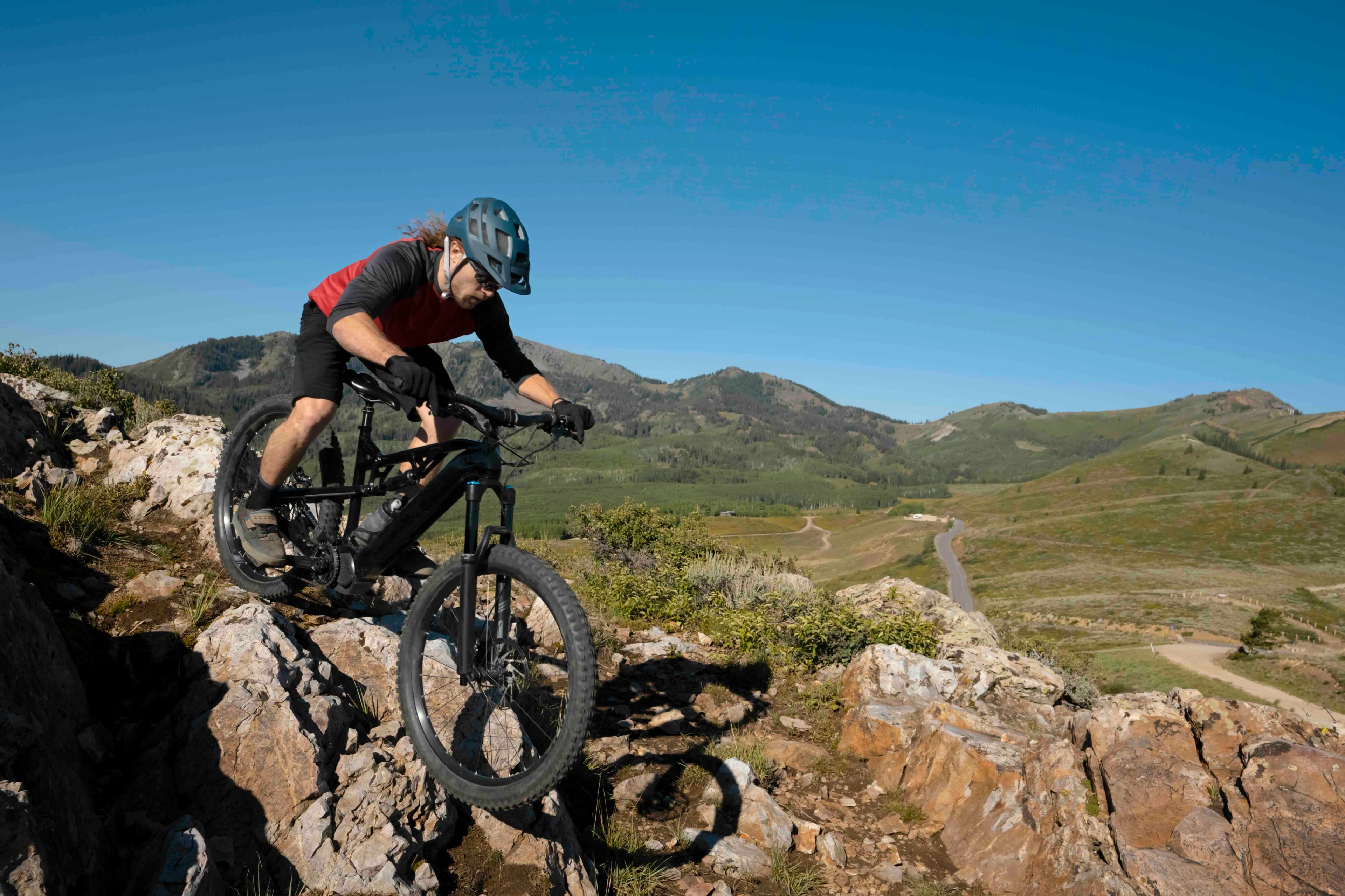 Une personne à vélo tout-terrain navigue sur un sentier rocheux par une journée ensoleillée, avec des collines verdoyantes et un ciel bleu clair en arrière-plan.