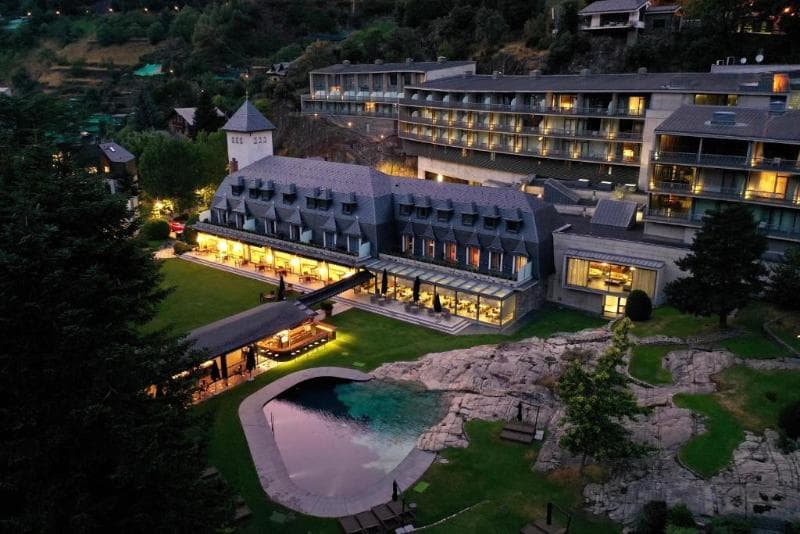 Vista aèria nocturna de l'Andorra Park Hotel, mostrant un complex de luxe il·luminat amb llums càlides. S'aprecia un edifici principal d'estil clàssic amb teulades de pissarra, juntament amb una estructura moderna integrada a la muntanya. En primer pla destaca una piscina de disseny orgànic envoltada de jardins verds, roques naturals i una zona de bar a l'aire lliure, tot sota l'atmosfera del capvespre en un entorn muntanyós.