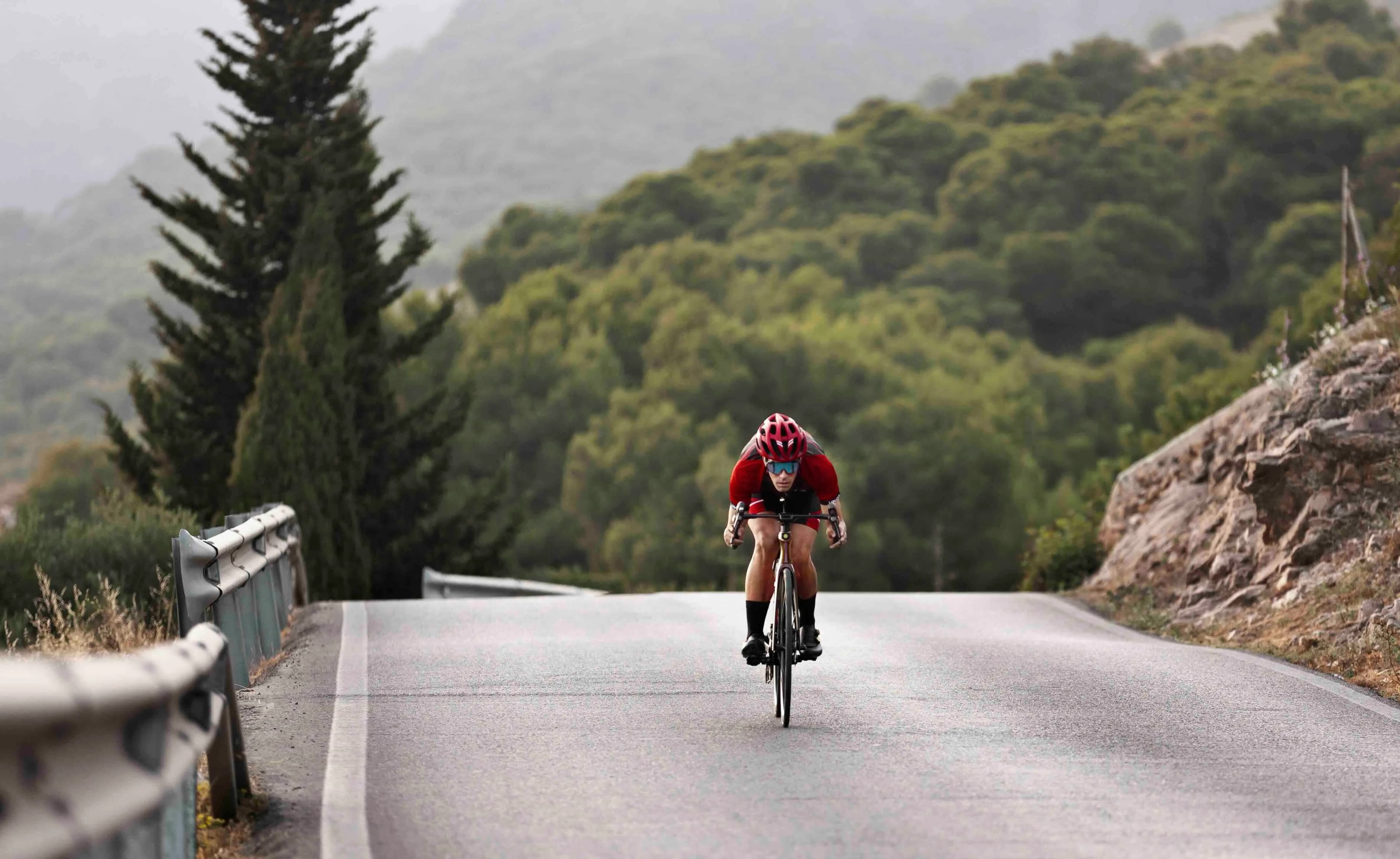 A cyclist in a red jersey and helmet rides a road bike uphill on a winding road, with green forested hills in the background.