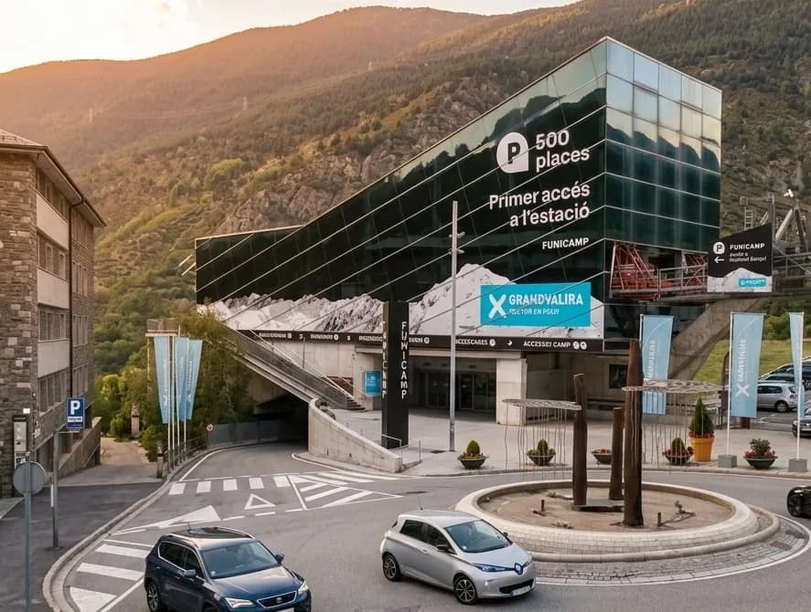 Un modern edifici de vidre, identificat com FUNICAMP i GRANDVALIRA, serveix com a entrada d'estació i aparcament al peu d'una muntanya boscosa, amb una carretera i cotxes en primer pla.