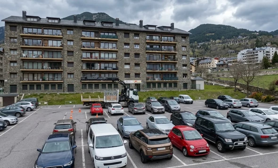 Un gran aparcament ple de diversos cotxes davant d'un edifici d'apartaments de pedra de diversos pisos. Al fons, turons verds i muntanyes s'eleven sota un cel ennuvolat, amb altres edificis visibles als vessants.