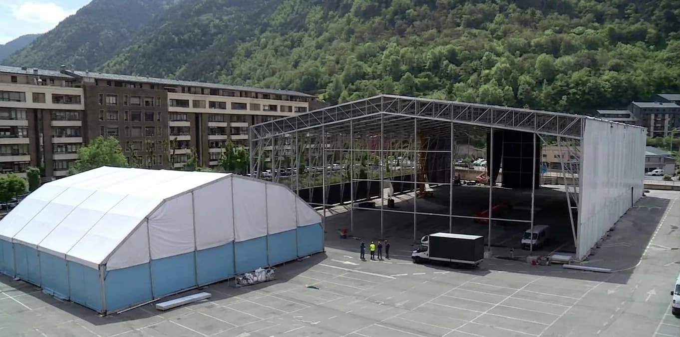 Vista aèria d'un gran aparcament amb dues estructures temporals per a esdeveniments: una carpa arquejada blanca i blava i una gran estructura metàl·lica oberta. Al fons hi ha edificis i muntanyes verdes.