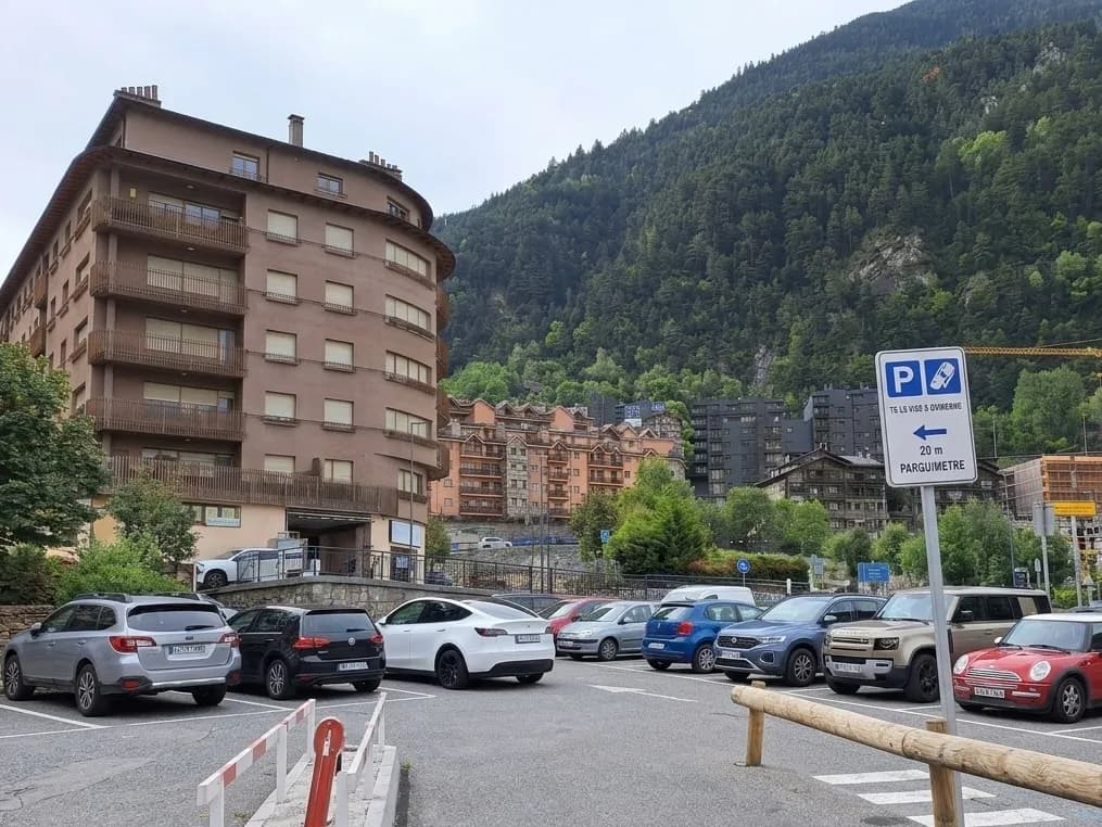 Un aparcament ple de diversos cotxes davant d'un gran edifici d'apartaments marró i corbat. Al fons, es veuen una muntanya boscosa i altres edificis, amb un senyal d'aparcament a la dreta.