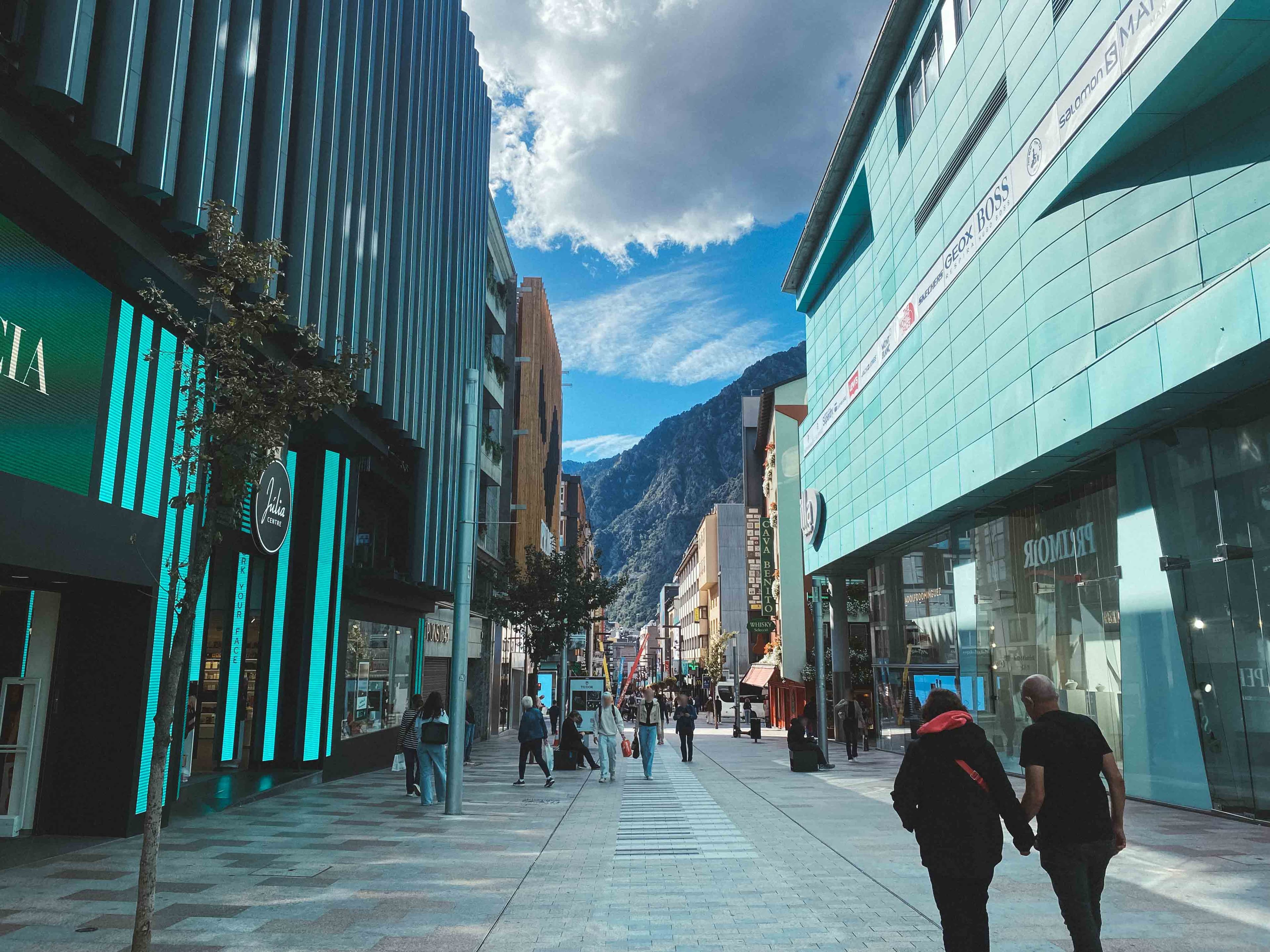 Una concurrida calle peatonal comercial con edificios modernos y escaparates, que se extiende hacia montañas bajo un cielo azul y nublado. Varias personas caminan por la calle, incluyendo una pareja en primer plano.