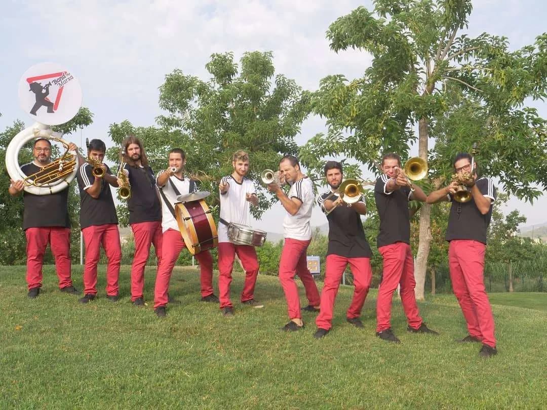 Una banda de nou homes amb samarretes negres i pantalons vermells posa sobre l'herba amb arbres de fons, sostenint diversos instruments com un sousafó, saxofons, tambors i trompetes. Alguns membres apunten els seus instruments cap endavant, i un sosté un cartell circular amb el logotip de la banda.