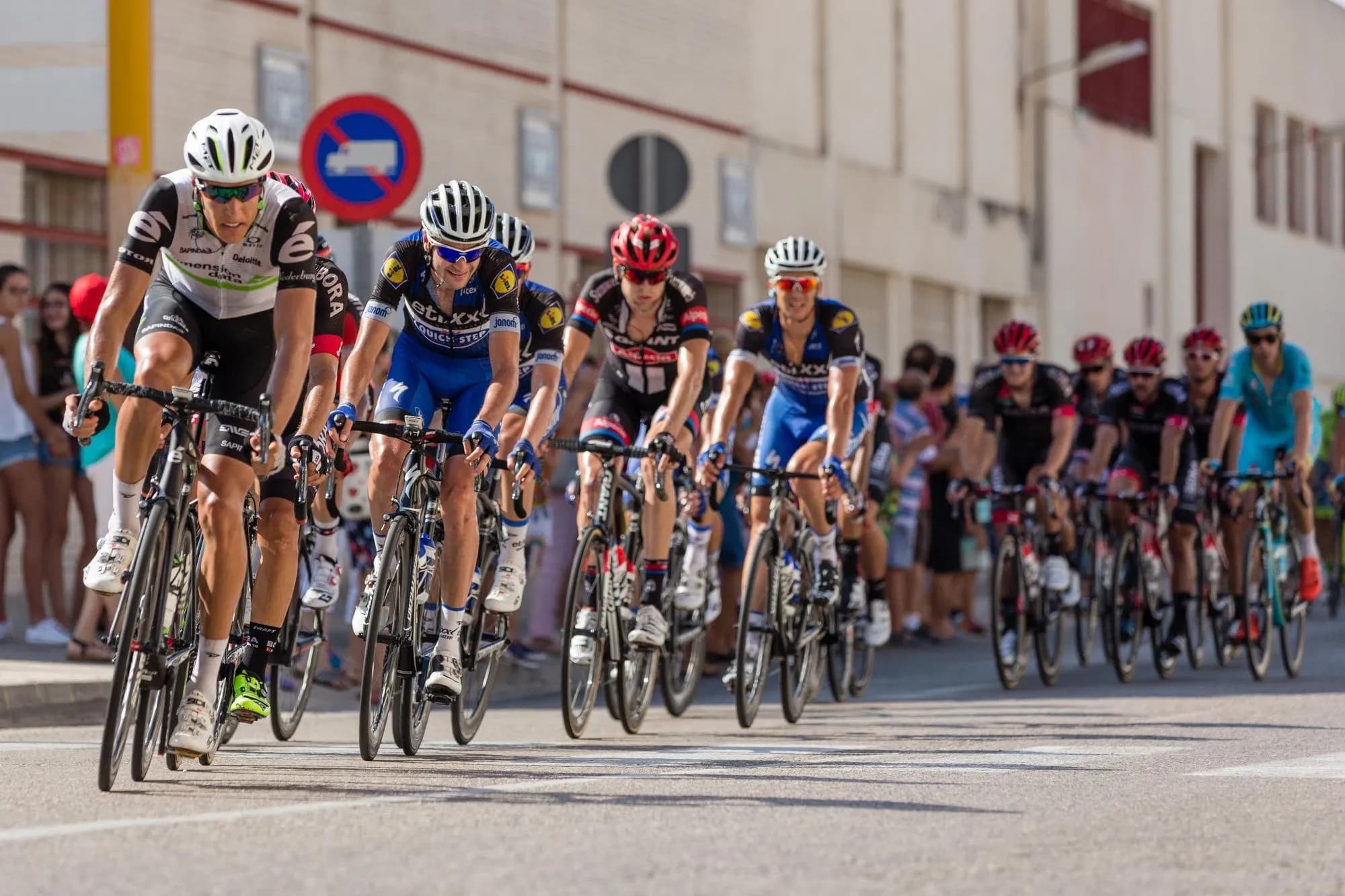 A group of professional cyclists in colorful jerseys and helmets race closely together on a sunny day, with spectators and buildings blurred in the background.