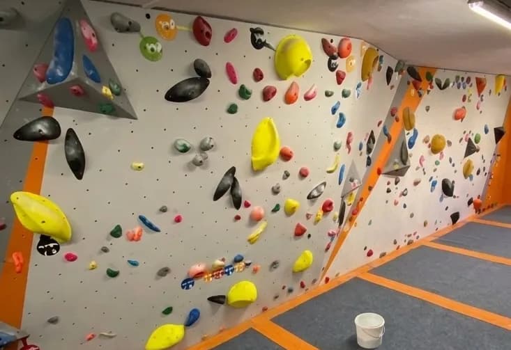 Un rocòdrom interior amb parets gris clar cobertes de nombroses preses d'escalada de colors de diverses formes i mides. El terra és gris fosc amb línies de seguretat taronges, i una galleda blanca es troba a la cantonada.