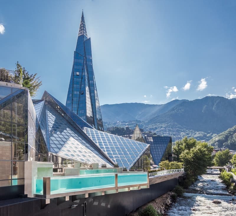 Un llamativo edificio moderno de cristal con una torre alta y puntiaguda y estructuras angulares de vidrio, que cuenta con una piscina infinita elevada con vistas a un río. Montañas verdes se elevan al fondo bajo un cielo azul claro.